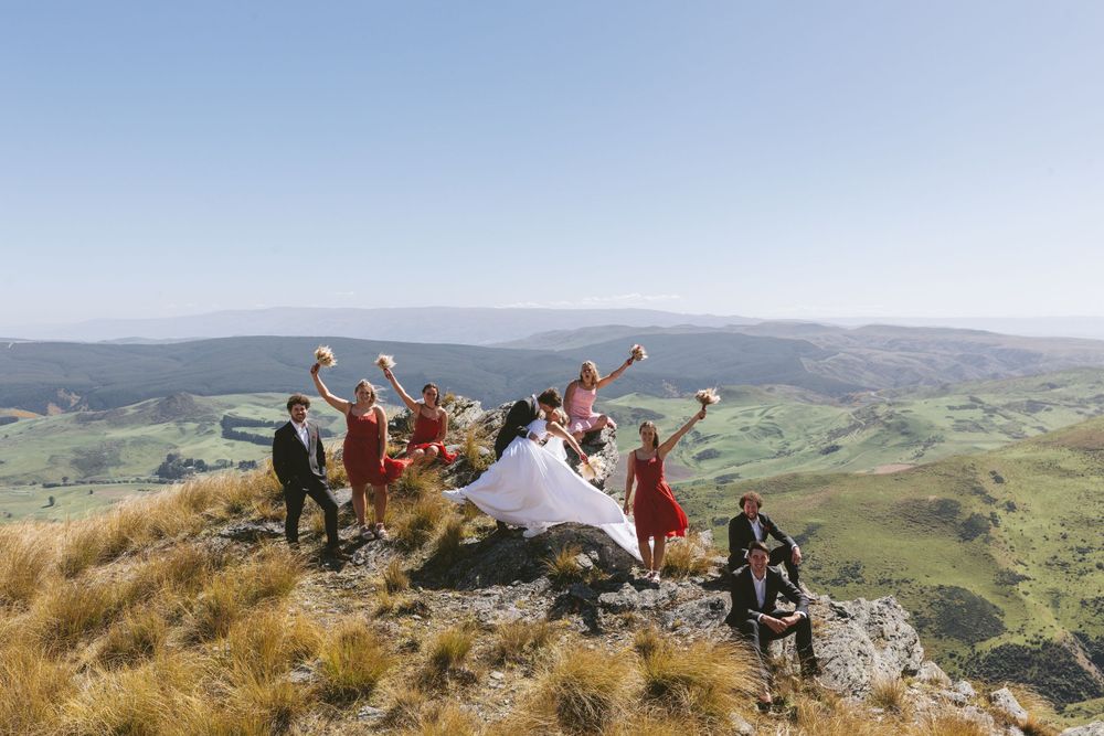 Joyful wedding party poses with raised arms on mountain summit overlooking vast landscape.