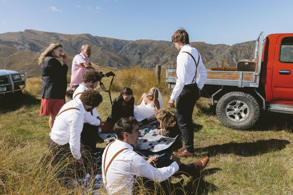 Wedding party gathered for picnic near red truck in mountain setting.