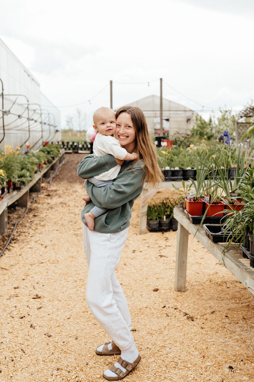 Someone wearing green sweatshirt and white pants walks through a plant nursery path with potted plants on both sides.