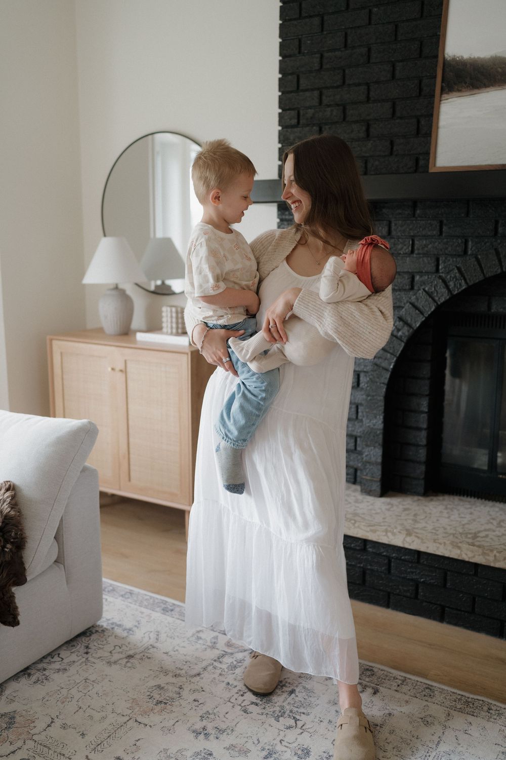 Parent and child standing by black brick fireplace in bright living room with white decor and furnishings.