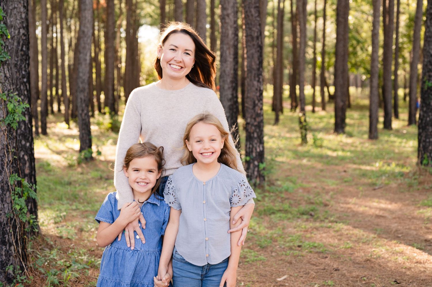 Family portrait of three people smiling in sunlit pine forest.