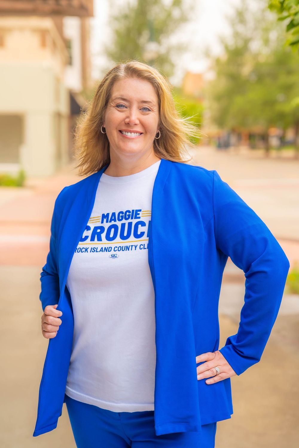 A person in bright blue blazer and campaign t-shirt stands smiling outdoors on a sunny day.