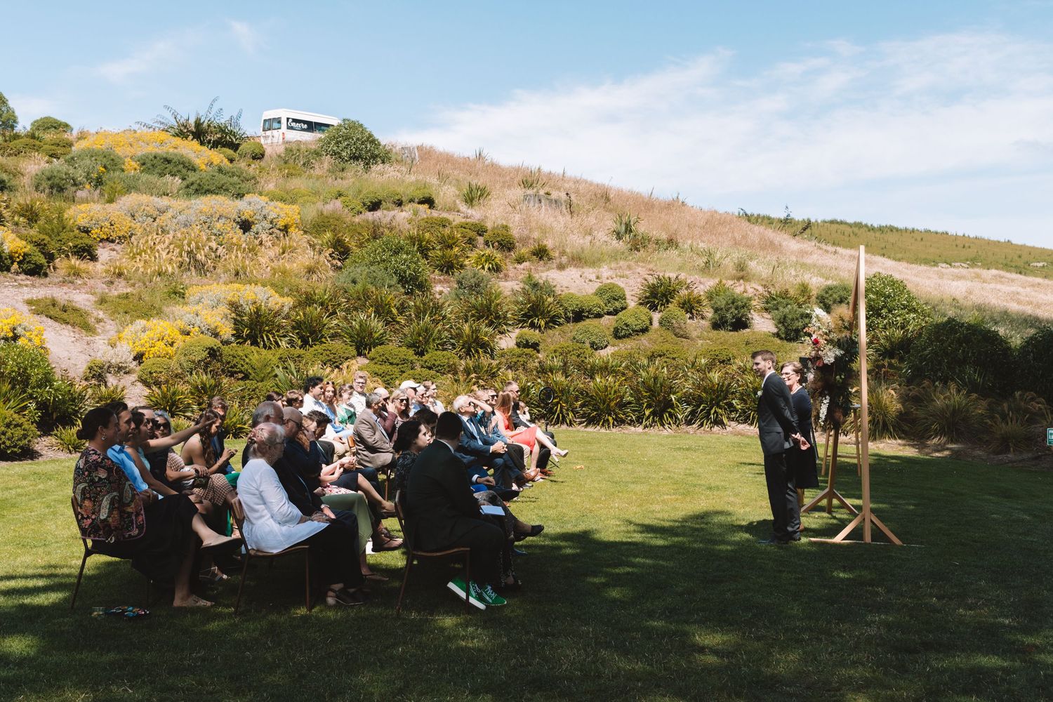 Wedding guests gathered on lawn seating listen to speaker during outdoor hillside ceremony.
