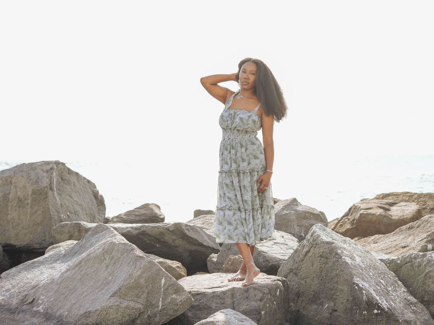 Woman in flowing dress poses on rocky beach landscape.