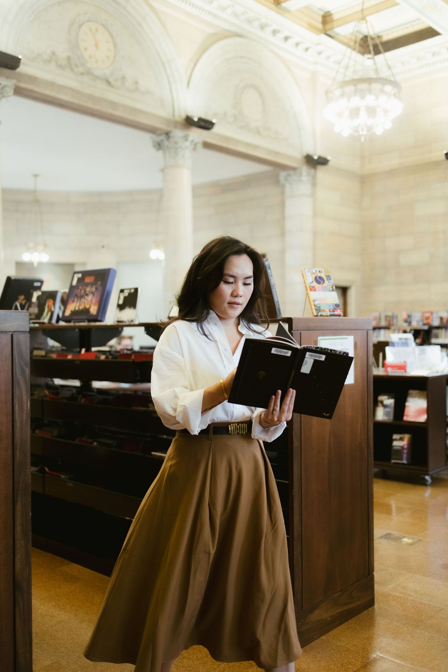 A person in a brown skirt reads intently while standing in an elegant library with ornate architectural details.