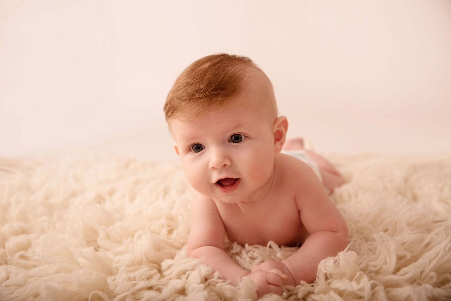 A happy baby lying on soft cream-colored fur during a professional photo session.