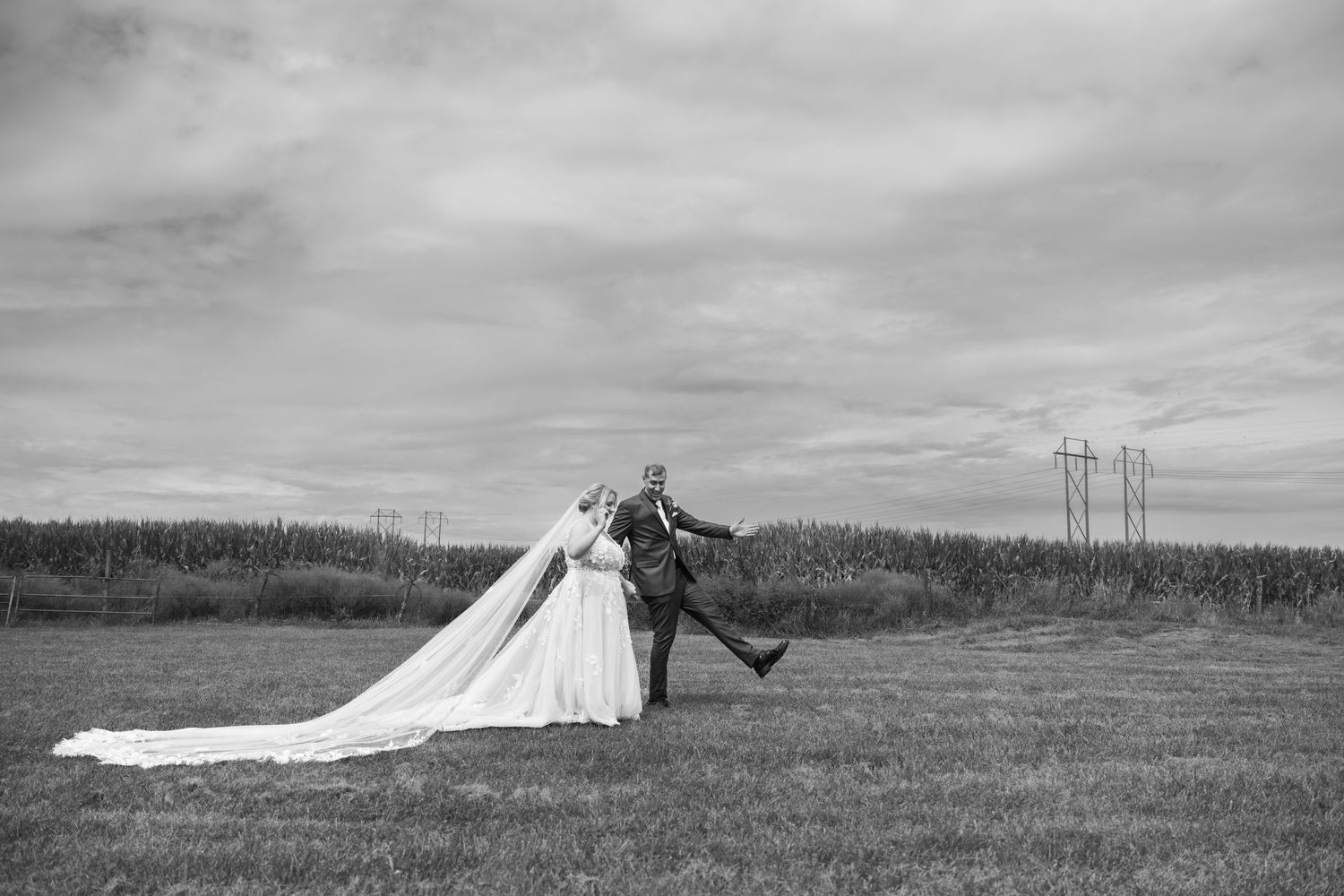 A playful black and white wedding photo in a field with power lines in the distance.