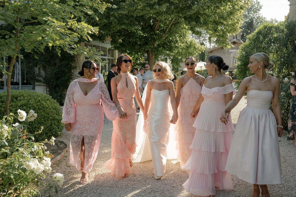 A sequence of wedding party members in pink and white dresses walking together through a garden path.