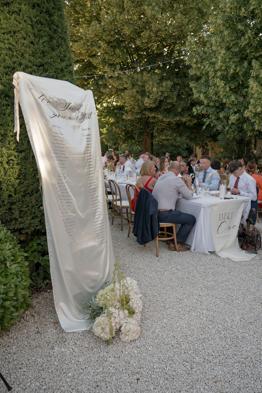 Outdoor wedding reception tables set up on gravel path with white tablecloths and chairs surrounded by greenery.