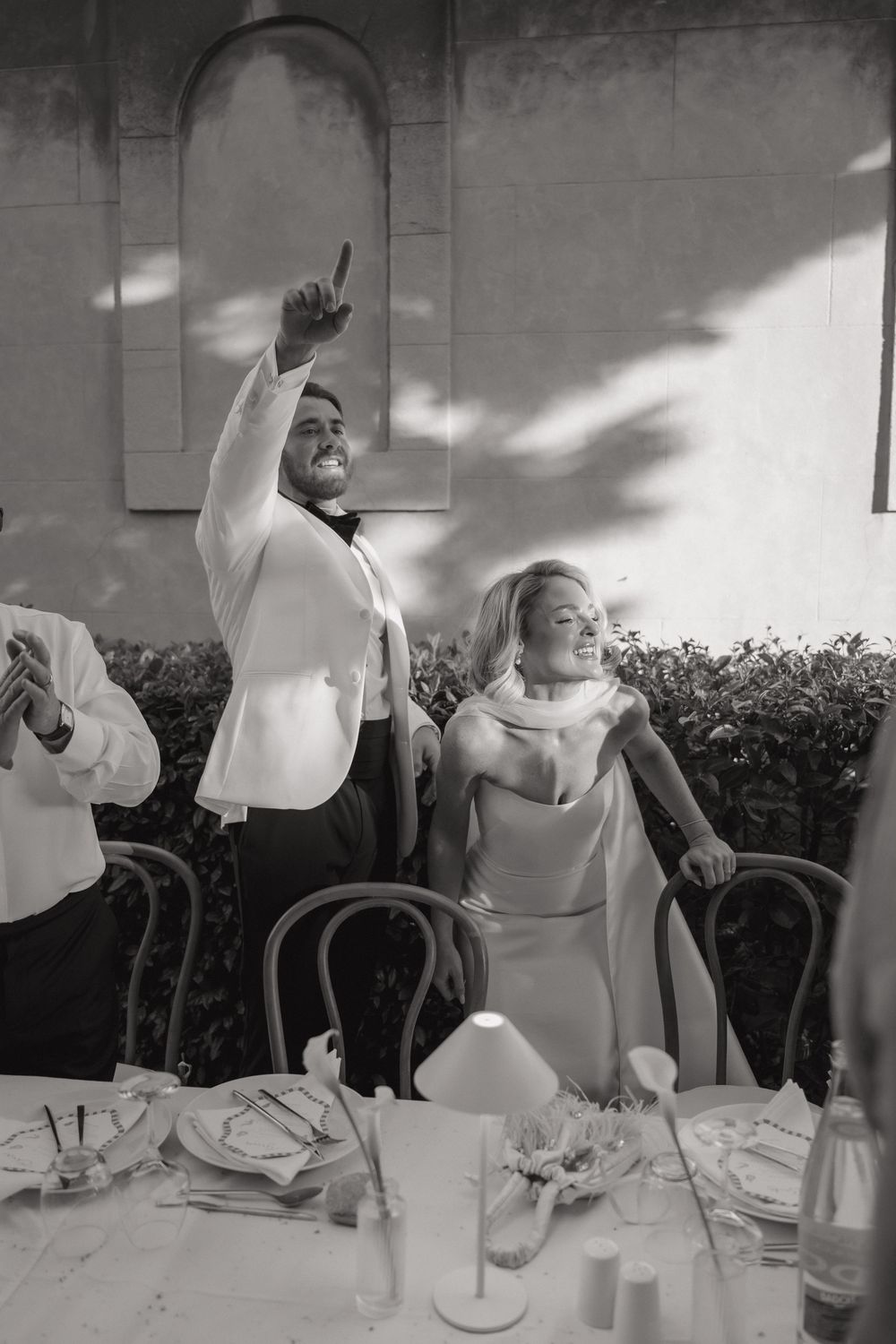 Black and white photo of two joyful people in white formal attire celebrating at an elegant reception dinner table.