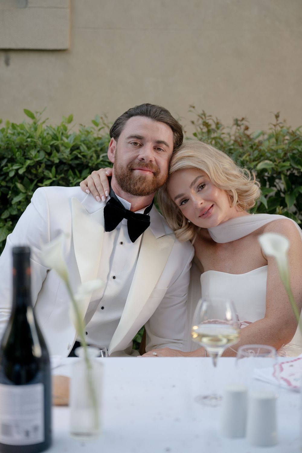 Happy couple dressed in formal wedding attire sit together at an outdoor reception table with wine glasses.