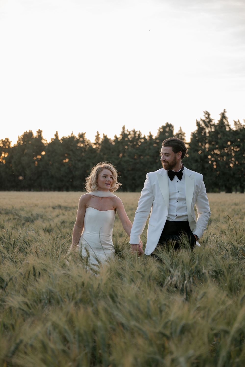 A couple walks together through a wheat field at sunset while wearing formal wedding attire.