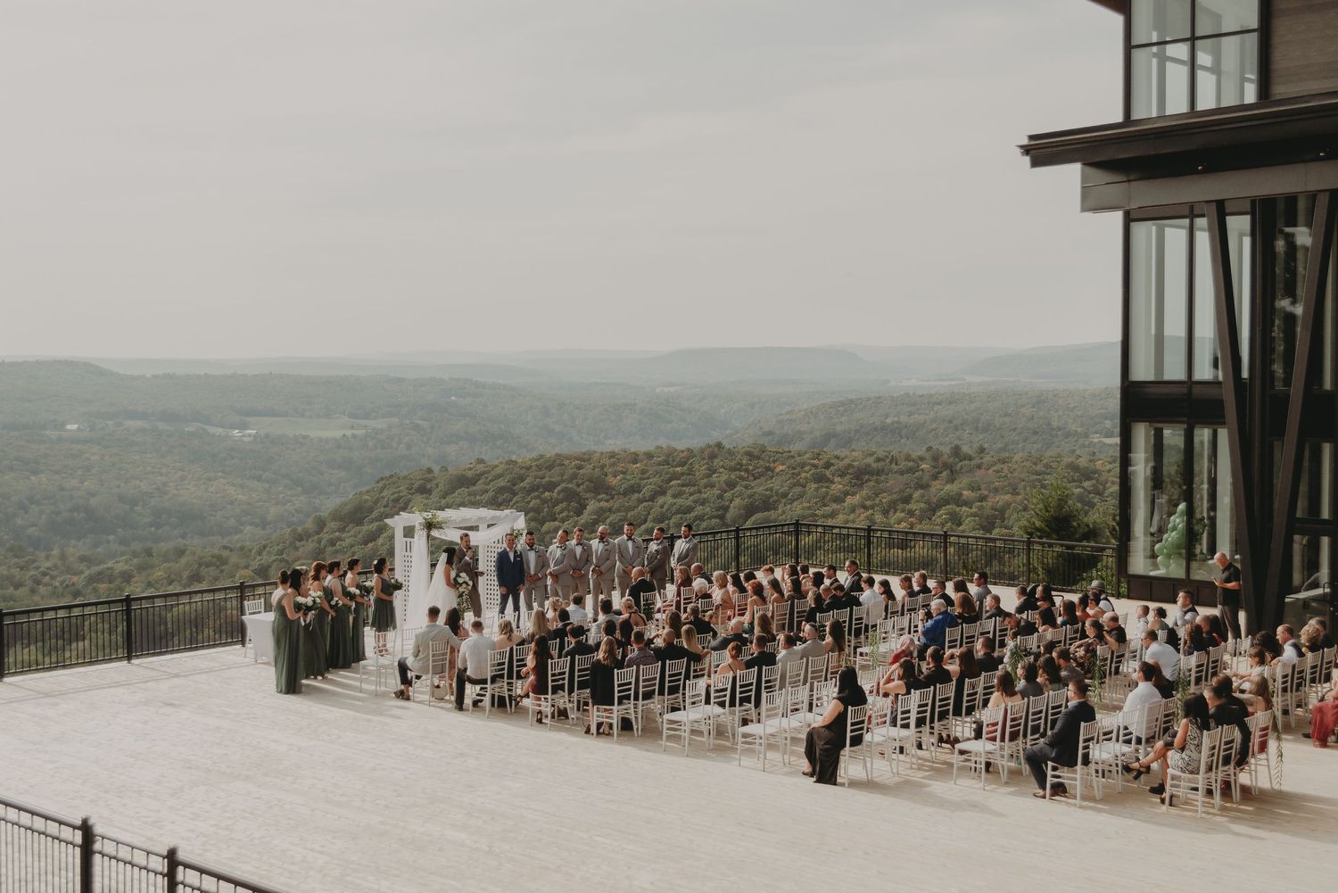 Panoramic view of outdoor mountain wedding ceremony with guests seated on elevated terrace.