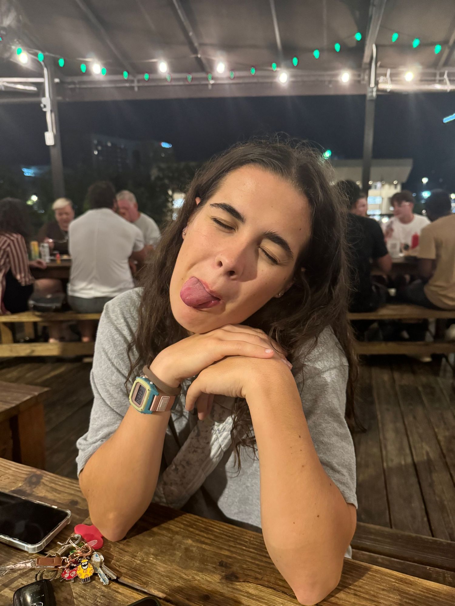 Playful person sticks out tongue while resting at a wooden table in an evening outdoor dining area with ambient lighting.
