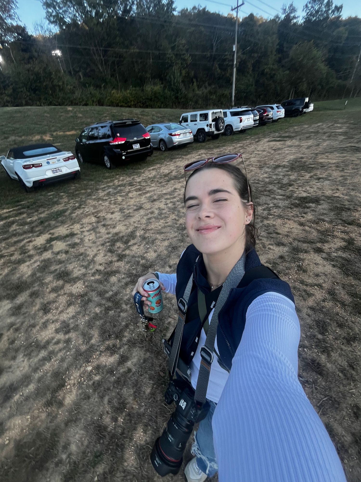 A photographer takes a selfie in outdoor setting with parked vehicles lined up in the background against wooded area.
