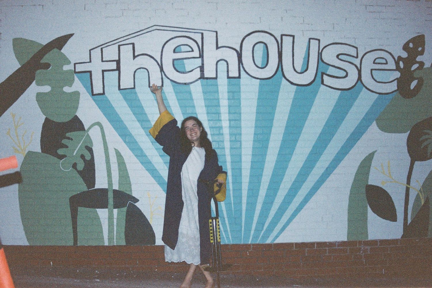 A person poses in front of a vibrant mural featuring The House text and silhouetted figures against a sunburst background.