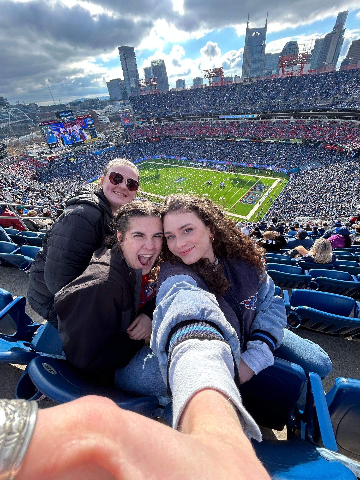 Aerial view of a packed NFL stadium with city skyline in background during daytime game. A group of three girls sitting in the stand smiling