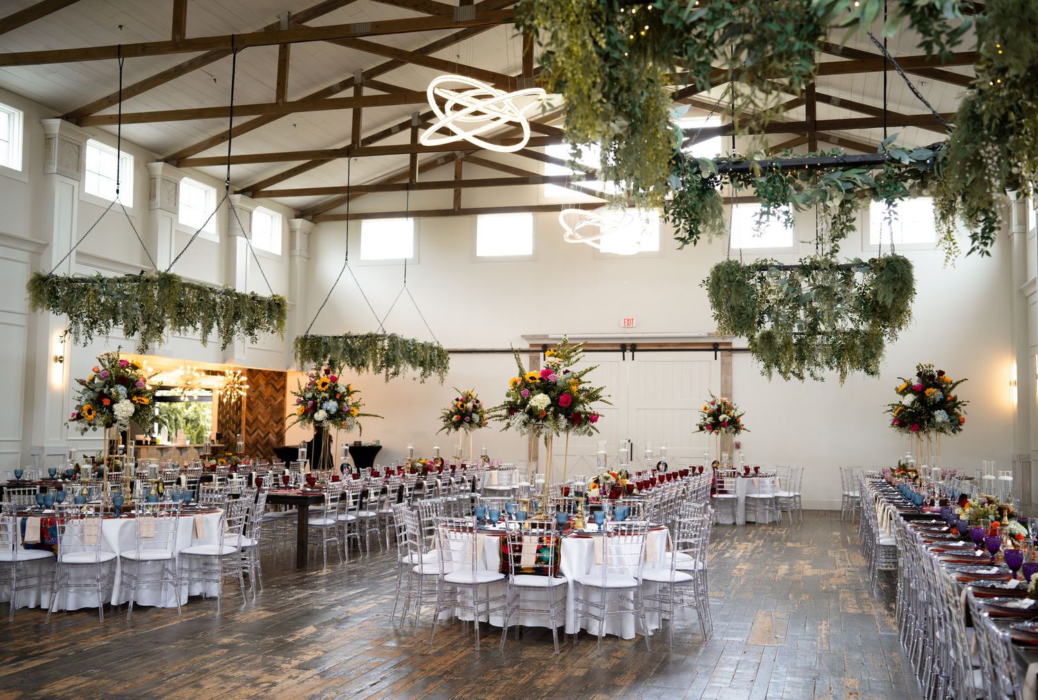 Elegant wedding reception hall with high ceilings, hanging greenery, and round tables set for a celebration.