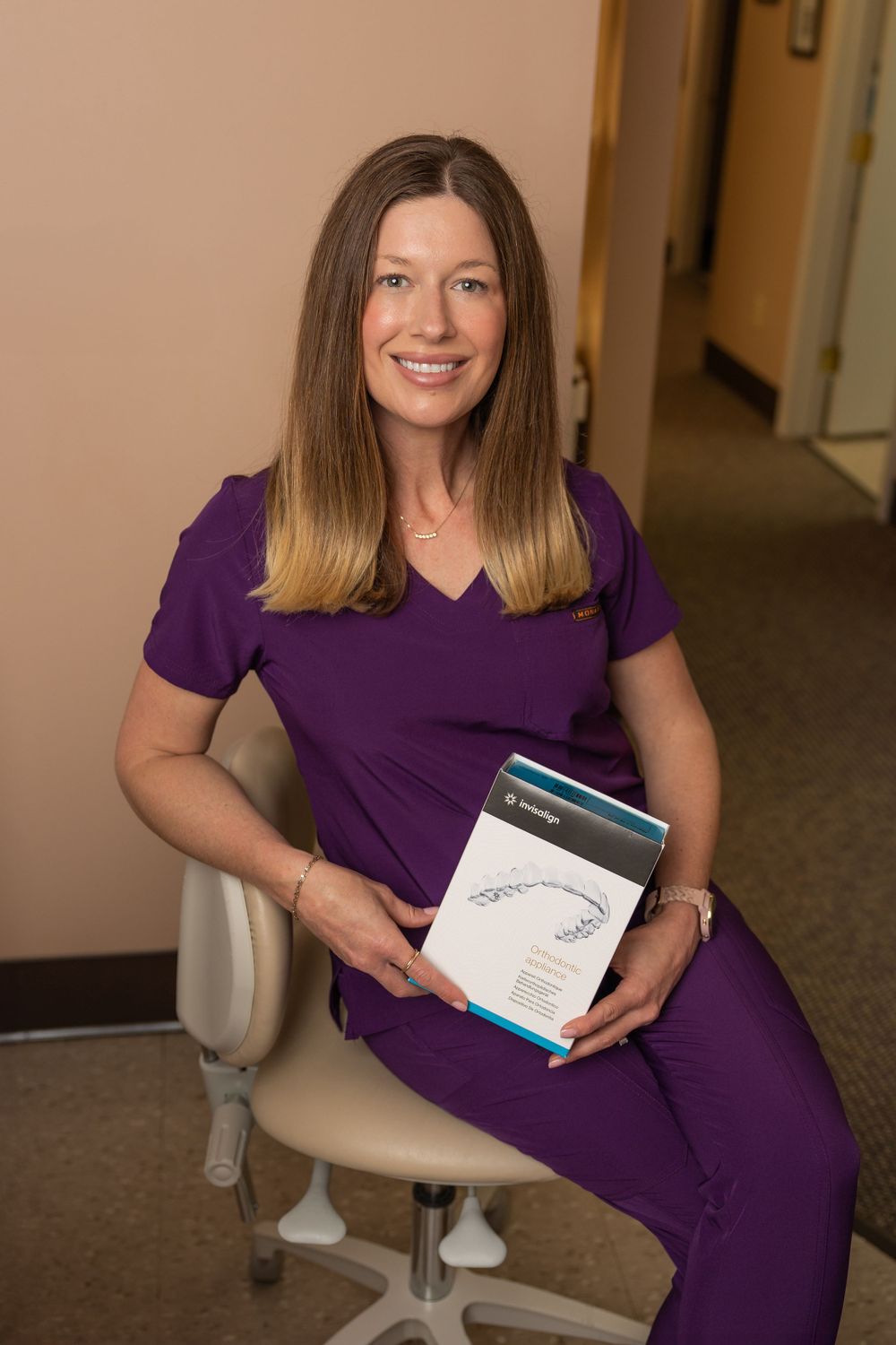 Healthcare worker in purple scrubs sits with Invisalign product guide in dental office.