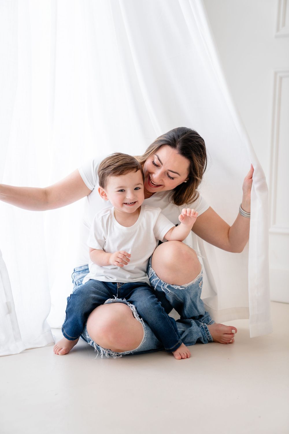 A pregnant mother and young child share playful moments together on a white backdrop in matching denim outfits.