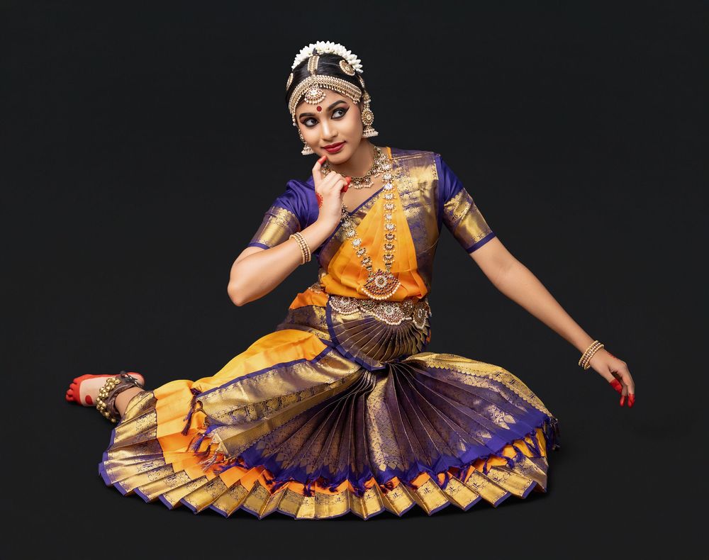 Classical Indian dancer performs in traditional yellow and purple Bharatanatyam costume with ornate jewelry against dark background.
