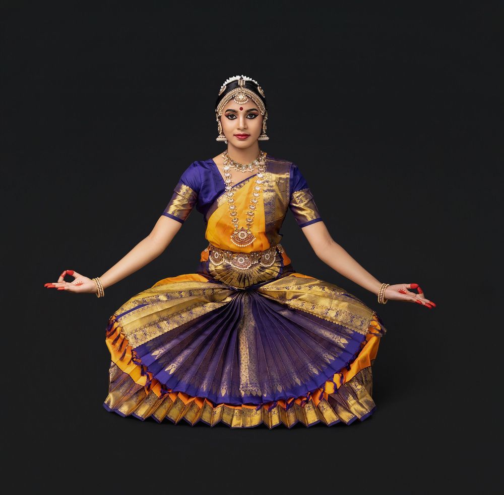 Classical Indian dancer in traditional yellow and purple costume performs elegant pose against dark background.