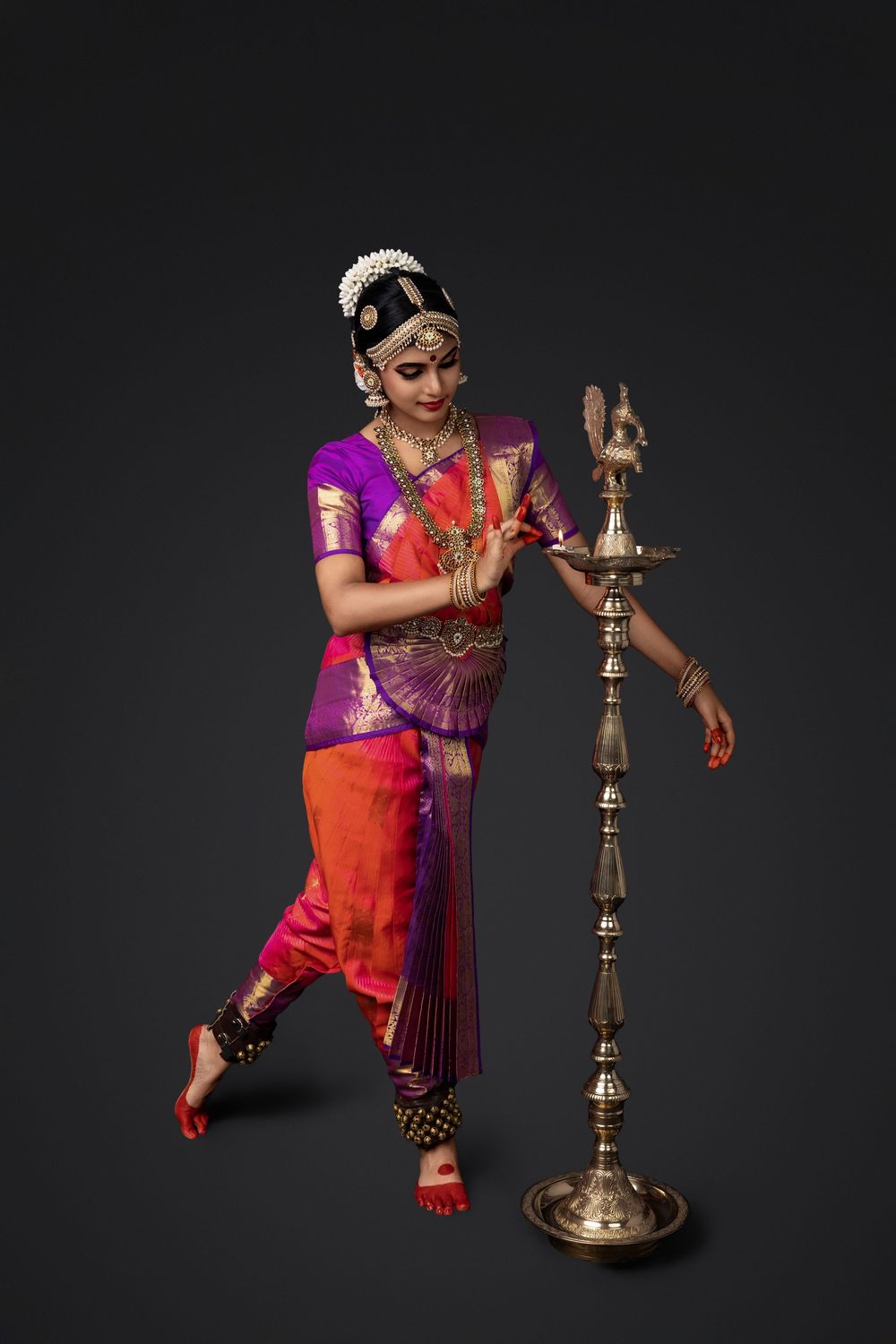 Classical Indian dancer in vibrant red and purple costume performs traditional moves beside an ornate brass lamp stand.