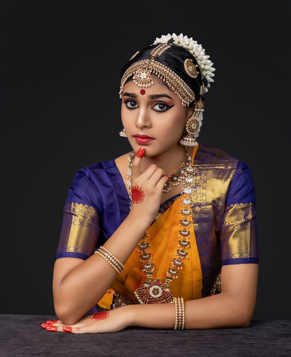 Classical Indian dancer in traditional orange and purple silk sari with ornate jewelry and makeup poses elegantly.
