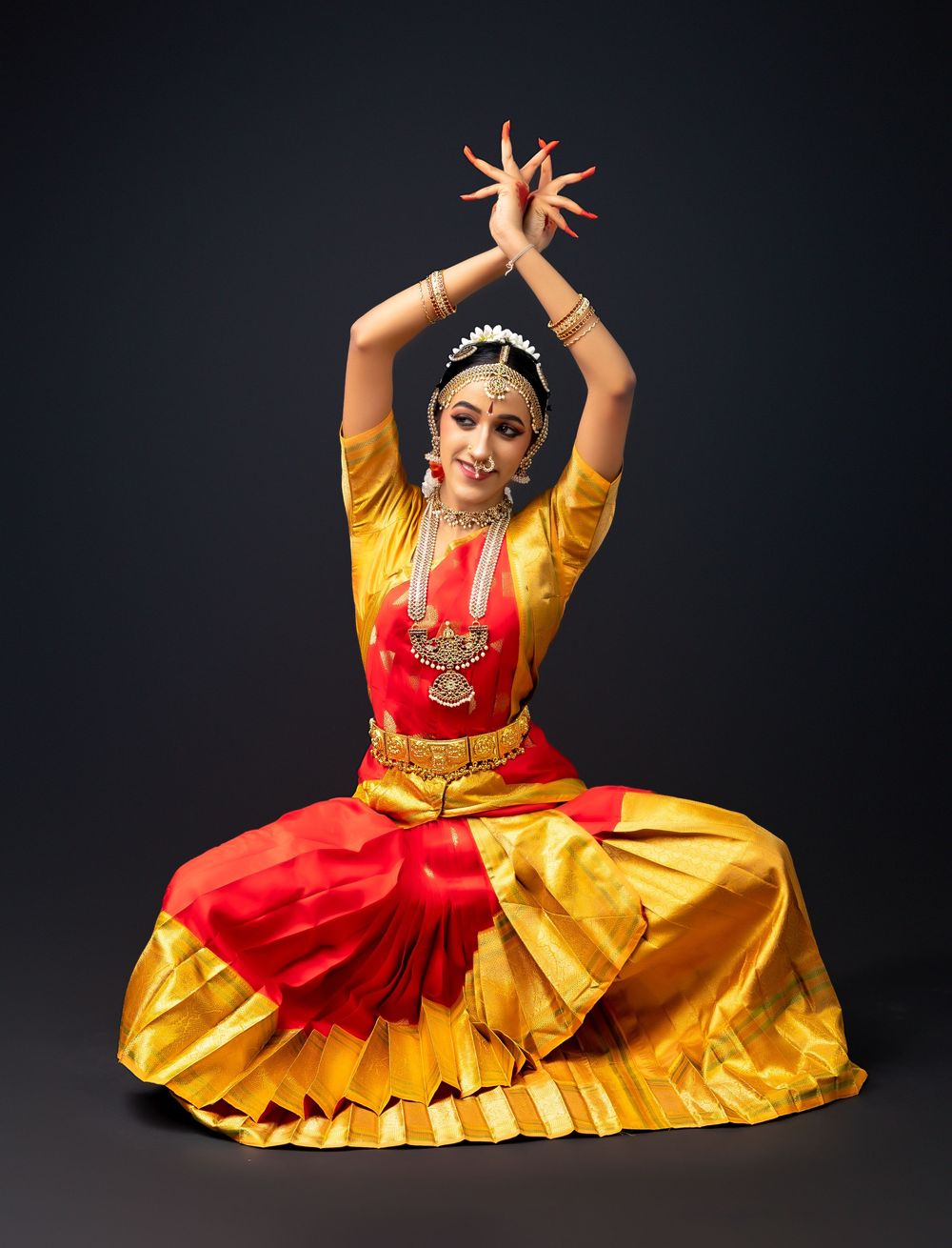 Traditional Bharatanatyam dancer in vibrant red and yellow costume performs seated mudra posture on dark backdrop.