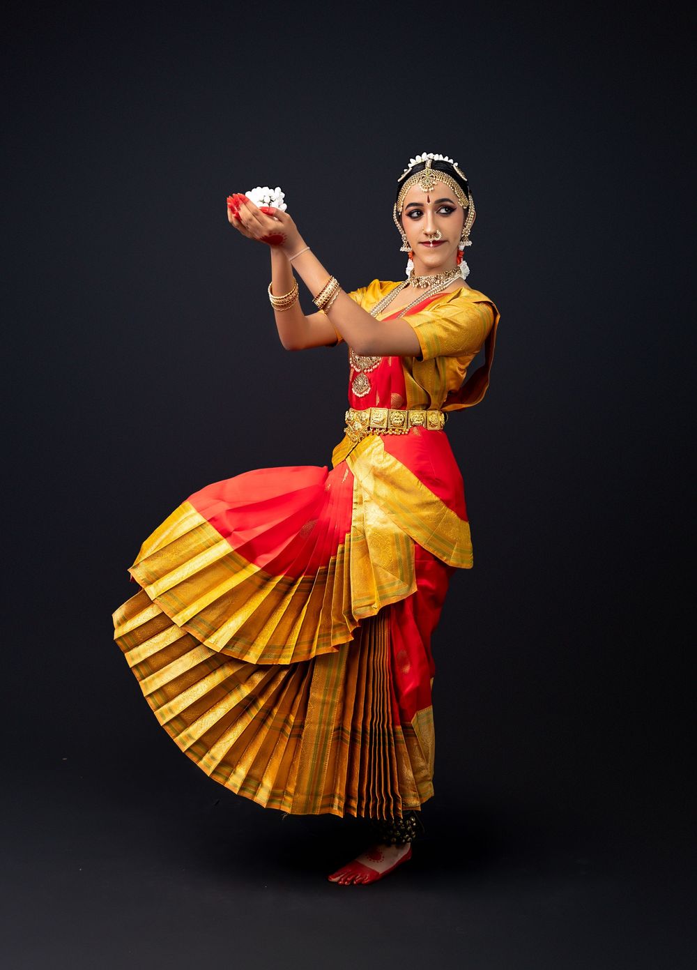 Classical Indian dancer performs Bharatanatyam in traditional red and gold silk costume against dark background.