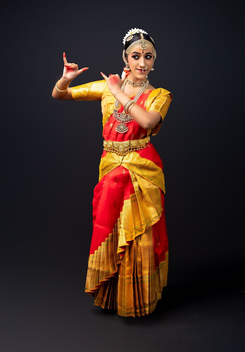Classical Indian dancer in vibrant red and yellow silk attire performs elegant Bharatanatyam dance movements against dark backdrop.