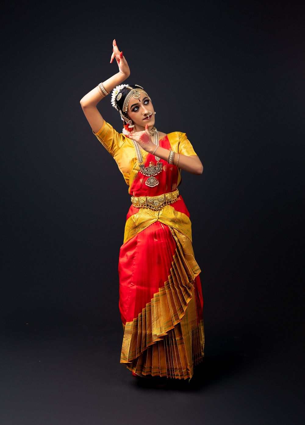 Classical Indian dancer performs in traditional red and gold silk costume with elegant arm positioning against dark background.