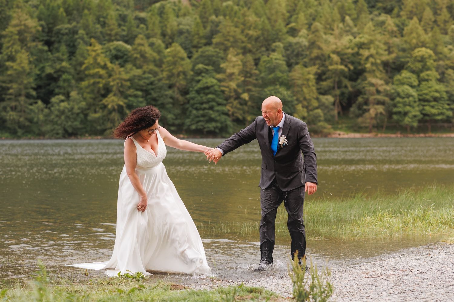 A couple in wedding attire holds hands and walks along a lakeside trail with evergreen trees in the background.