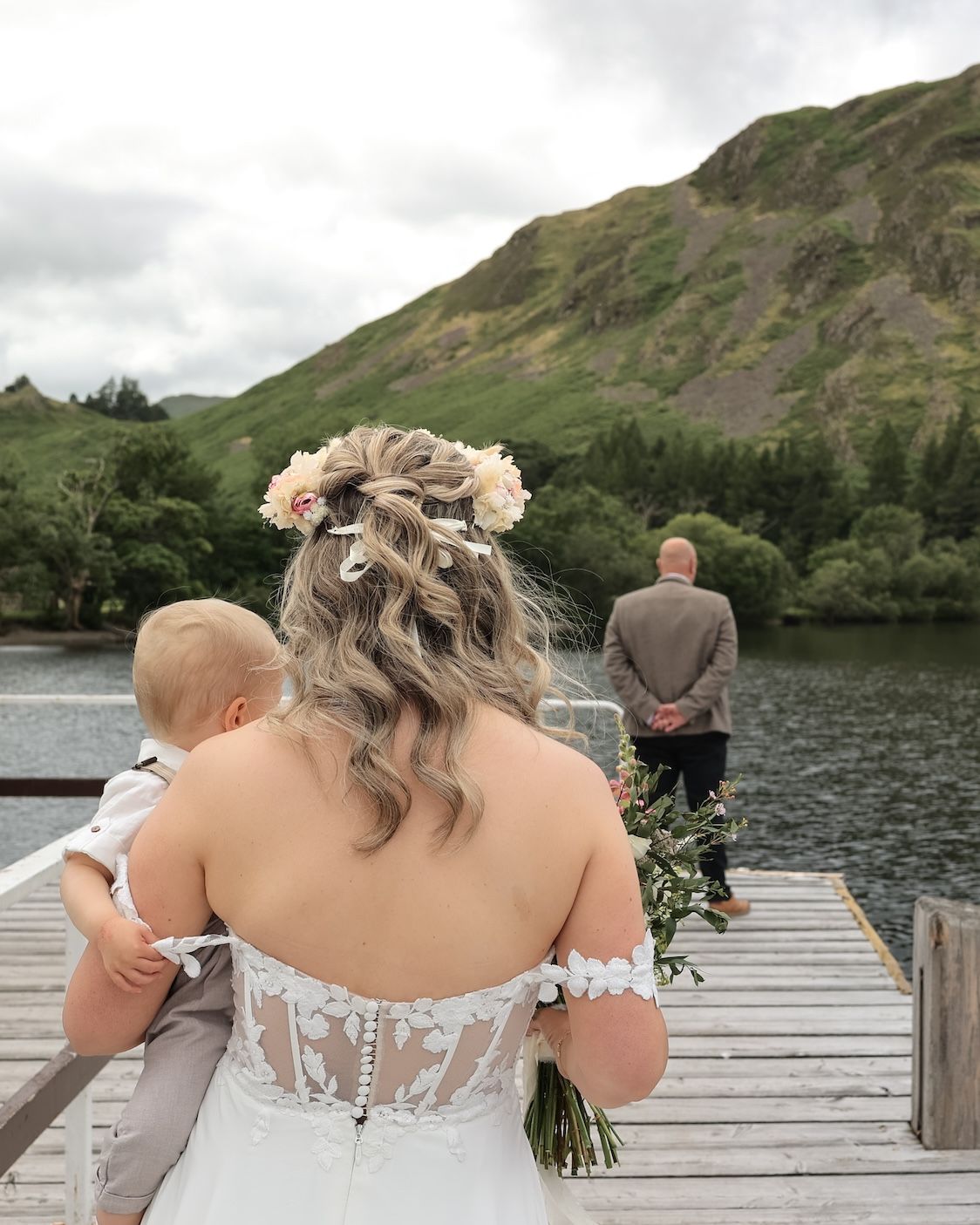Wedding couple stands at the edge of a wooden dock overlooking a serene lake surrounded by green mountains.