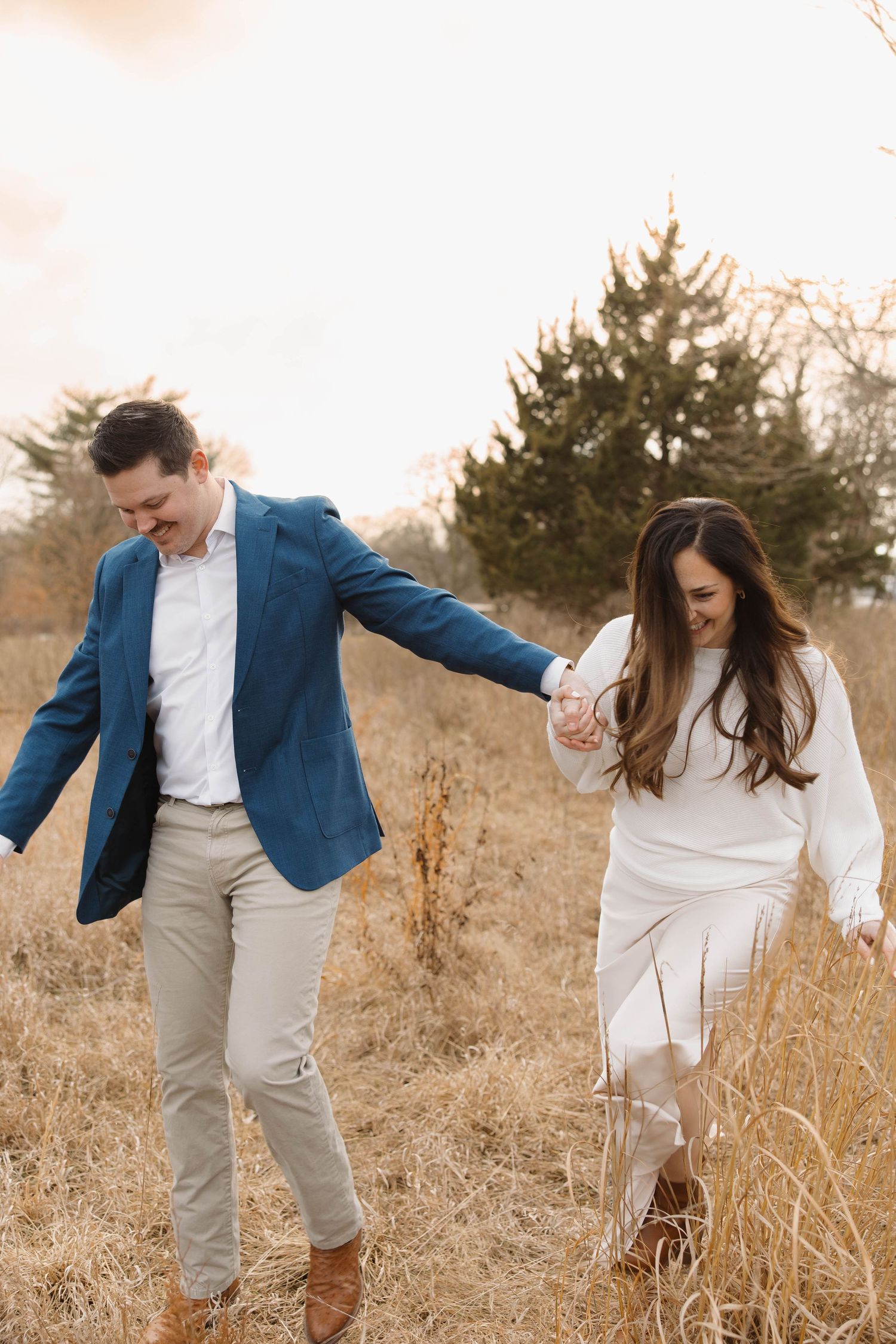 A couple in coordinating blue and white outfits walks hand-in-hand through tall grass in an autumn field.