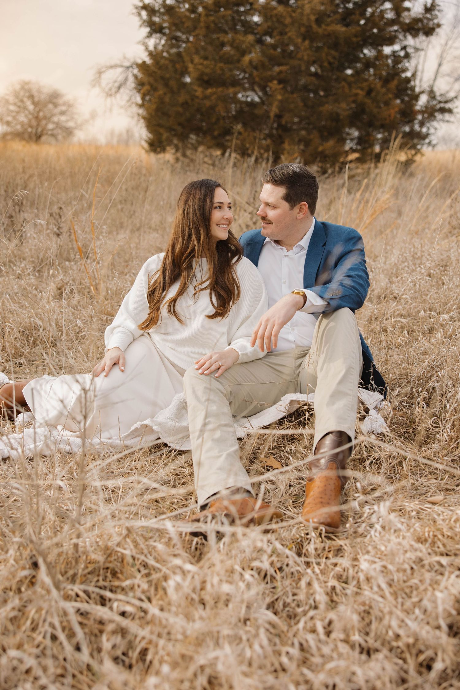 A romantic couple sits together on a blanket in a golden field with evergreen trees in the background.