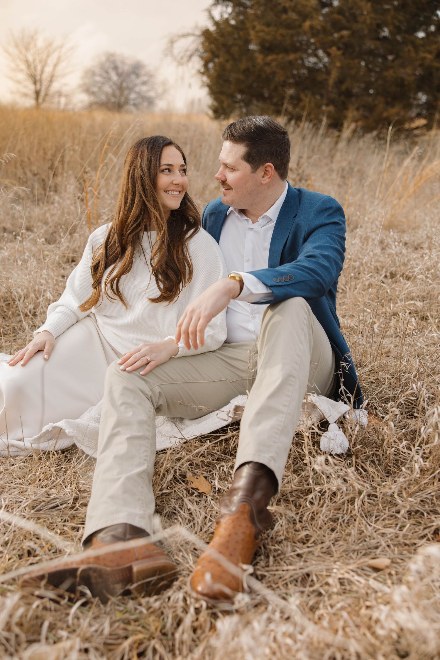 A couple wearing white and blue sits intimately in a golden fall field as sunlight filters through dry grass.