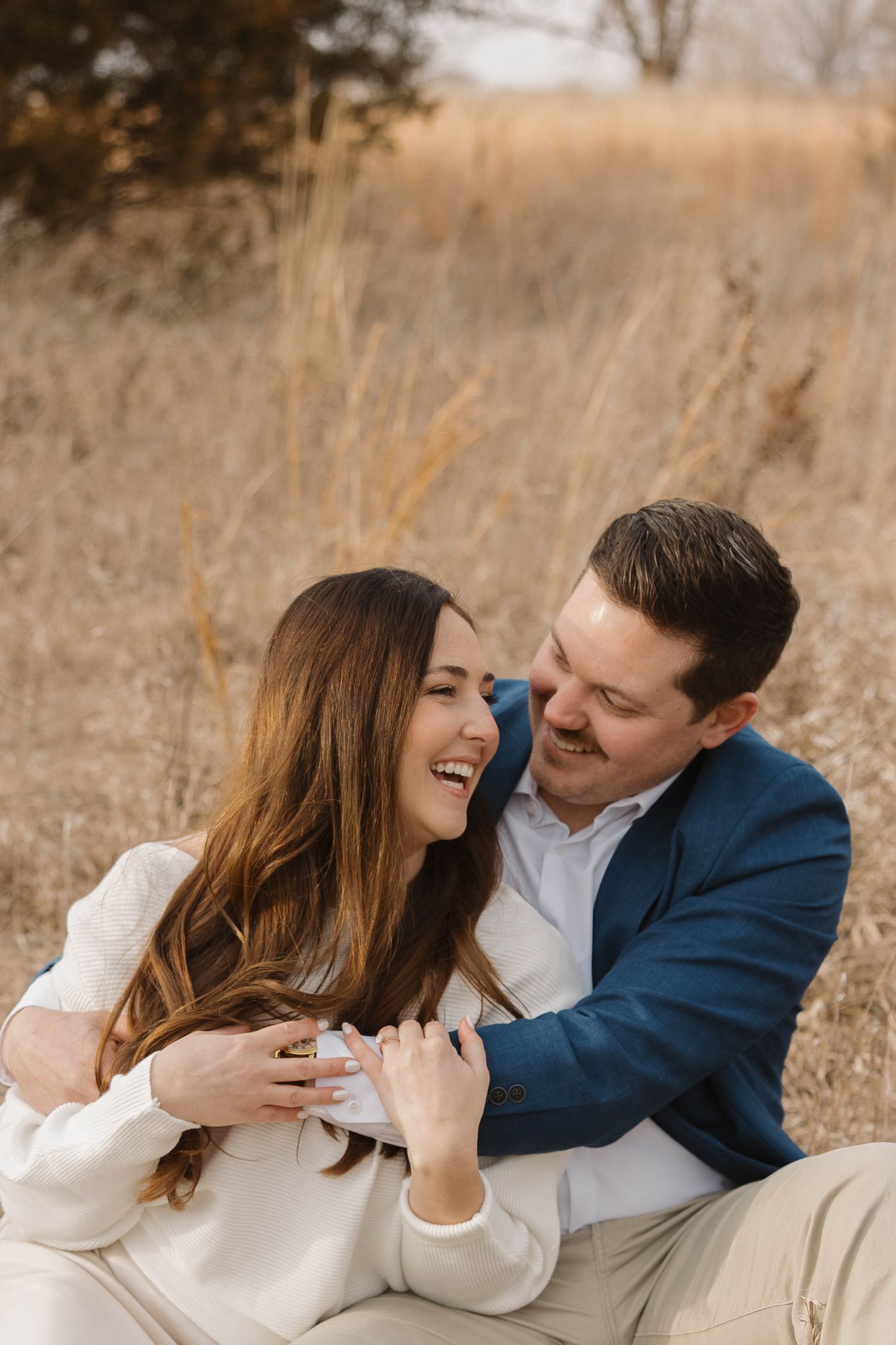 A couple shares a joyful moment together while sitting in tall autumn grass during an outdoor photo session.