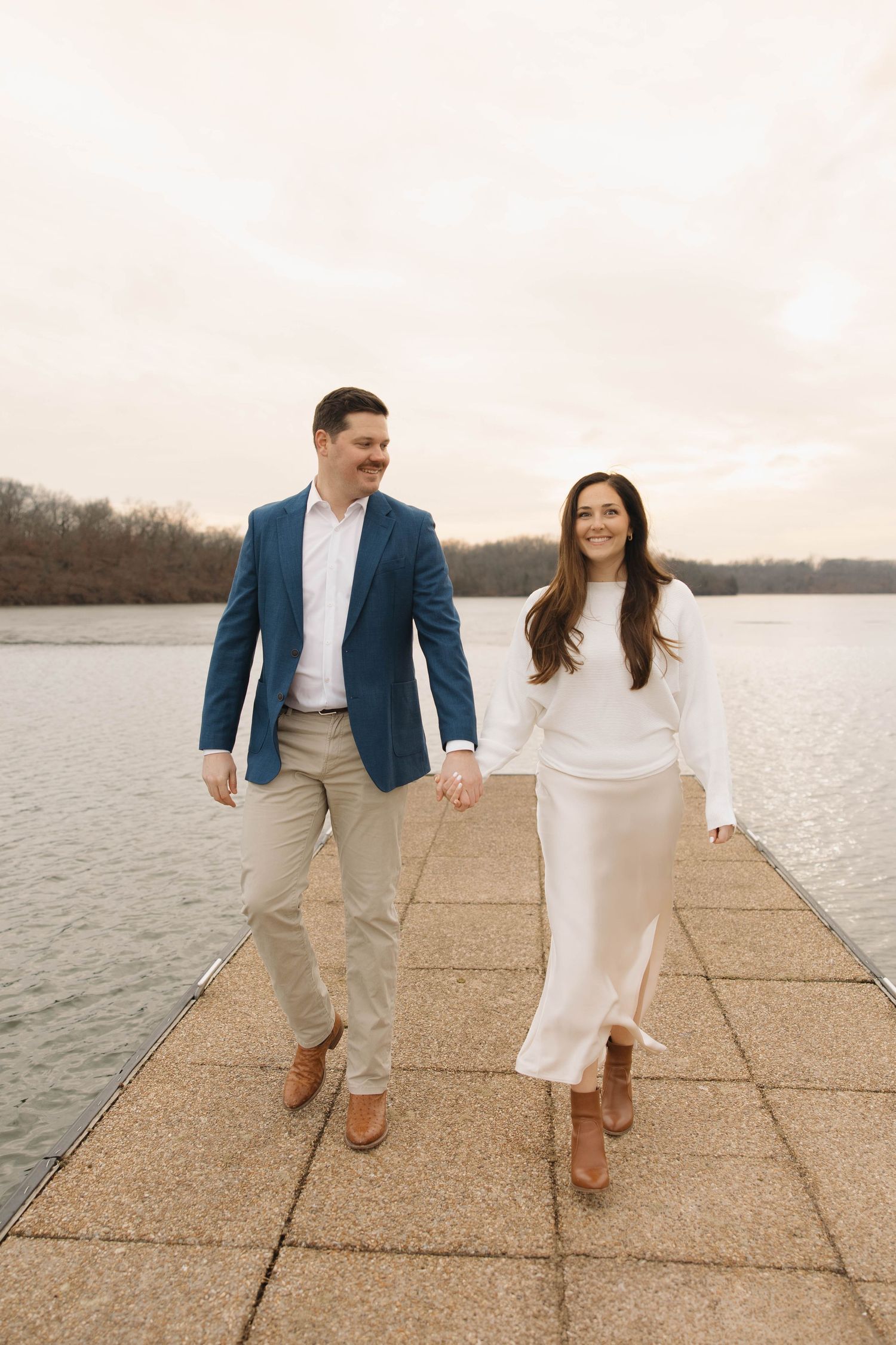 Couple walking and holding hands along a waterfront concrete path wearing blue blazer and white dress.
