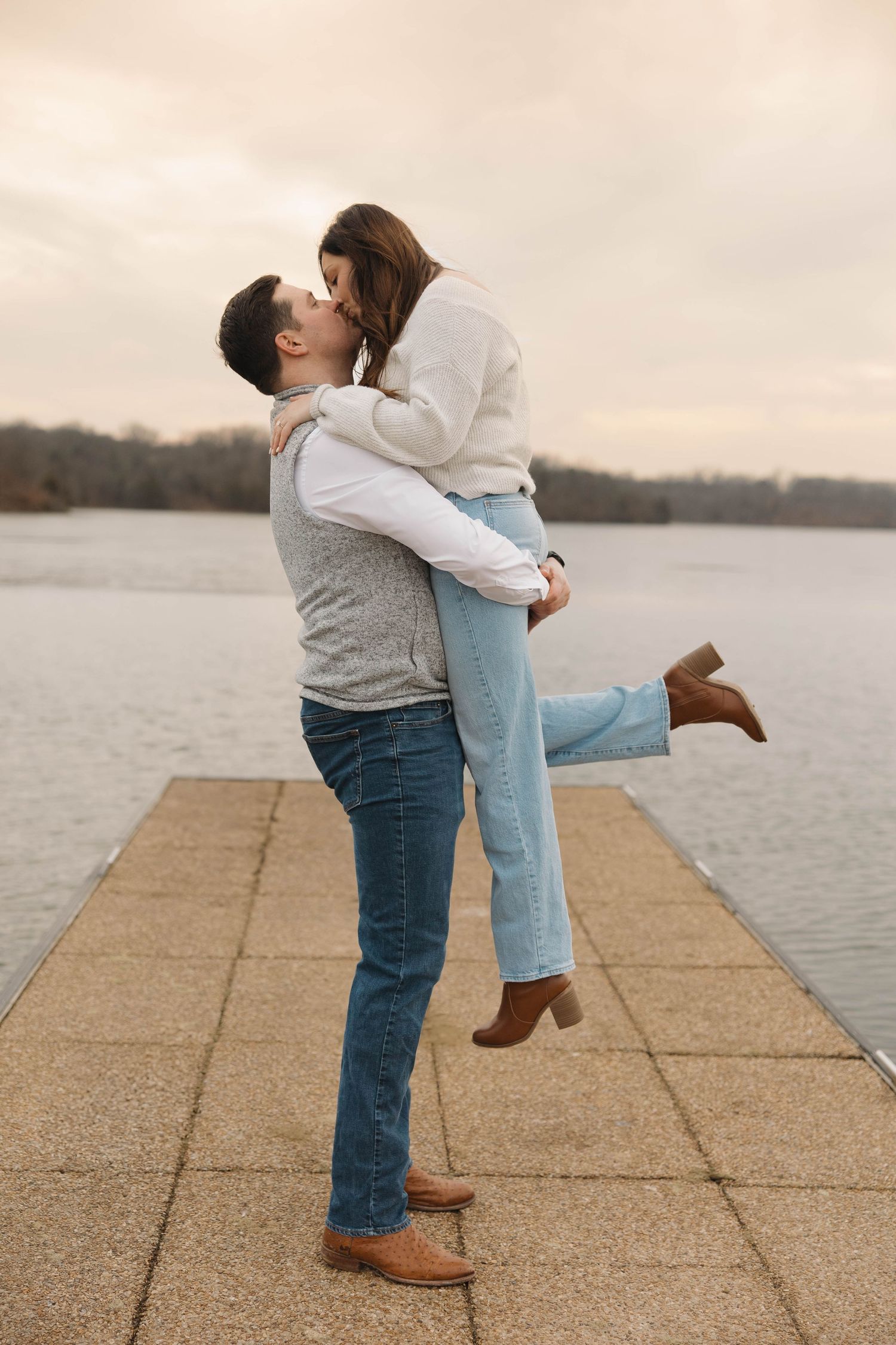 A couple shares a romantic moment while embracing on a concrete pier by a serene waterfront.