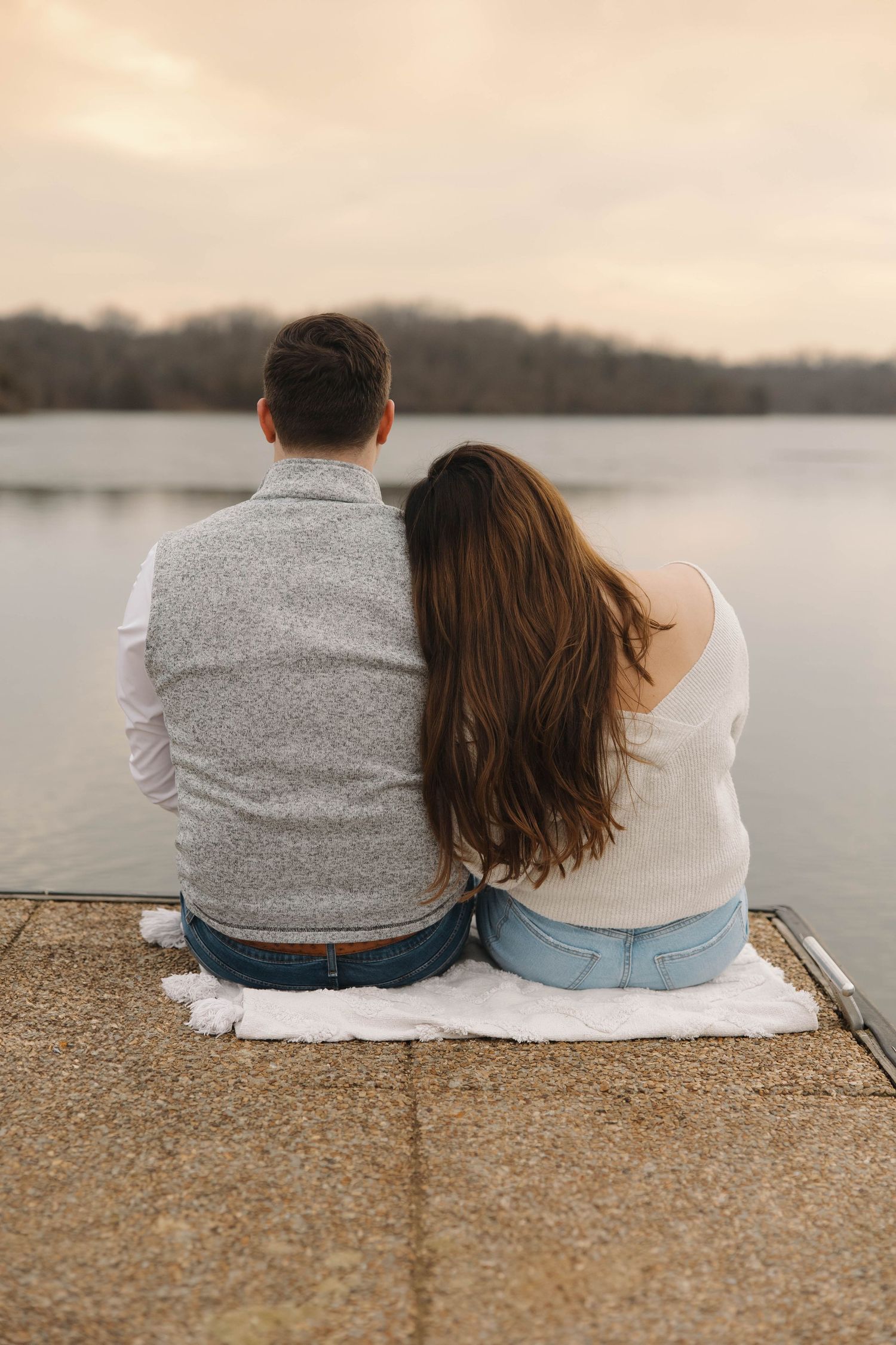 A couple sit close together on a shoreline while watching a serene lake at sunset.