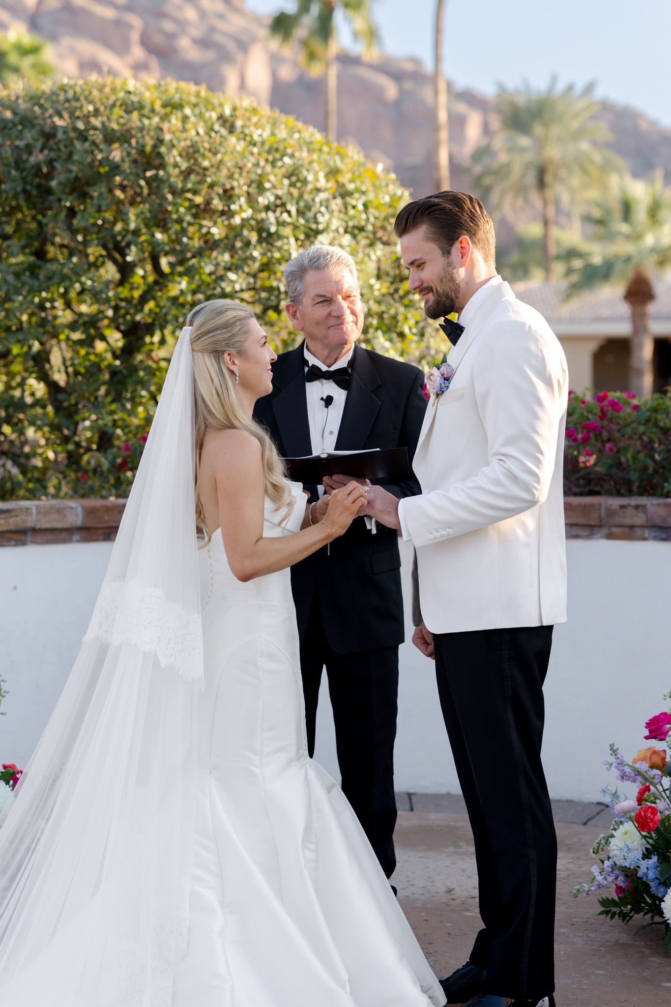 Couple exchanges vows during an outdoor wedding ceremony at a desert resort with palm trees and mountains in background.
