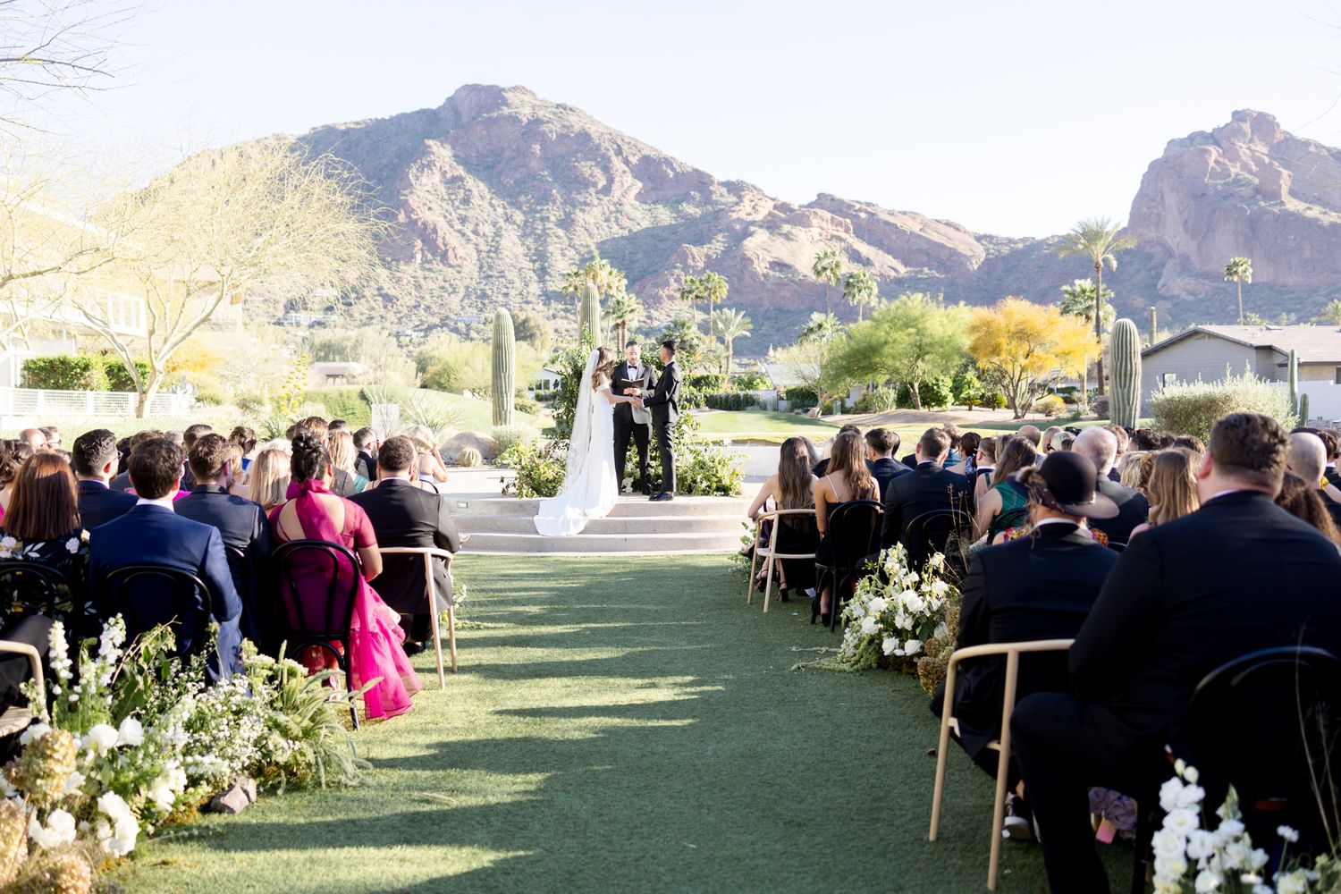Outdoor wedding ceremony at a scenic desert venue with mountain backdrop and green lawn surrounded by desert flora and guests.