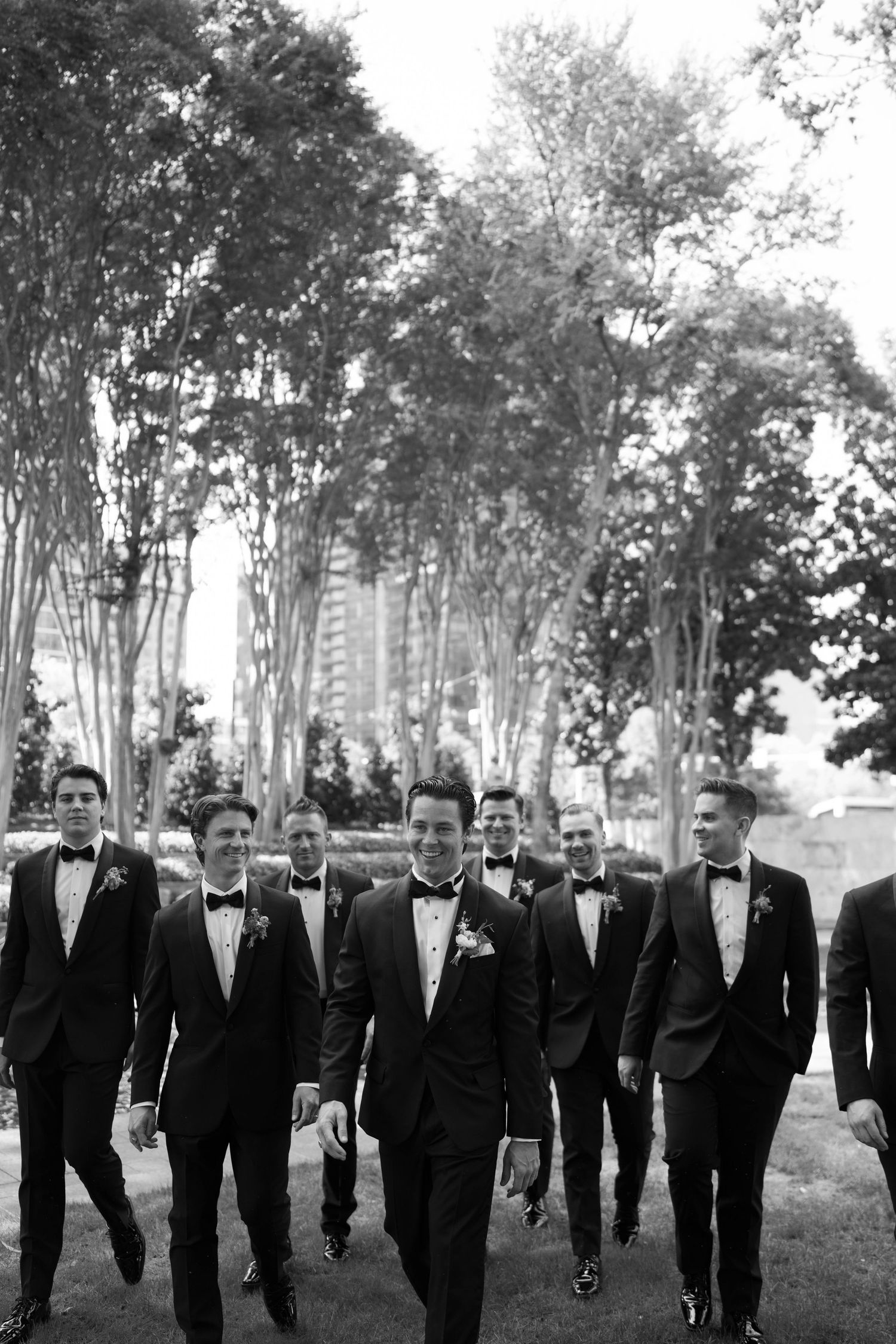 Groomsmen in black tuxedos and bow ties walk together through a tree-lined path in elegant black and white photo.