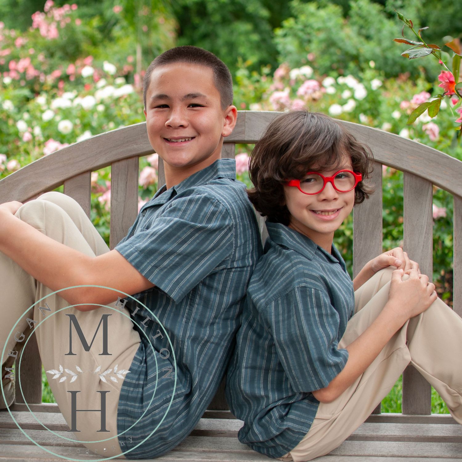 Two kids sitting back-to-back on a wooden garden bench with pink and white flowers blooming in the background.