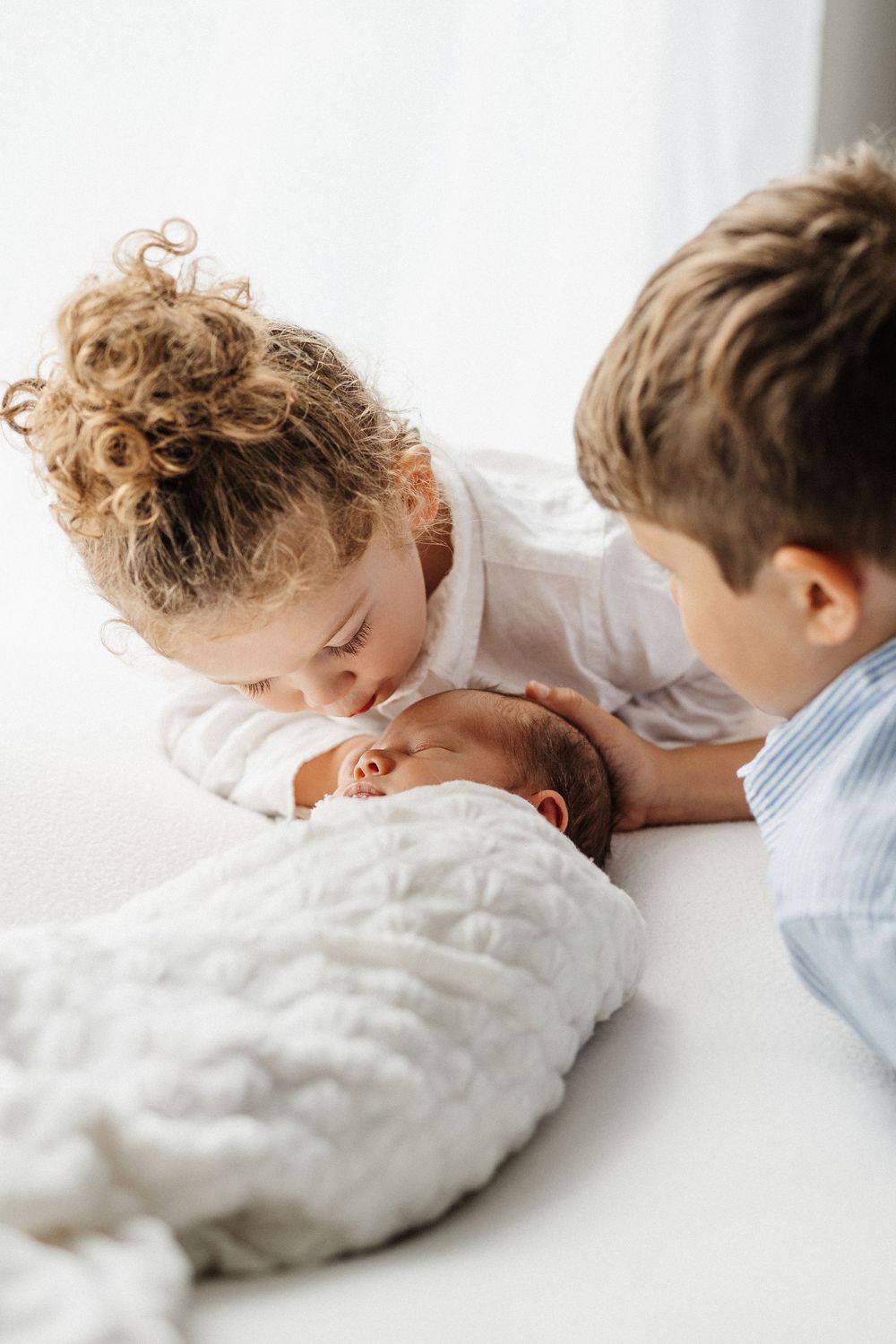 Two young children laying closely together on white bedding, showing a tender sibling moment.