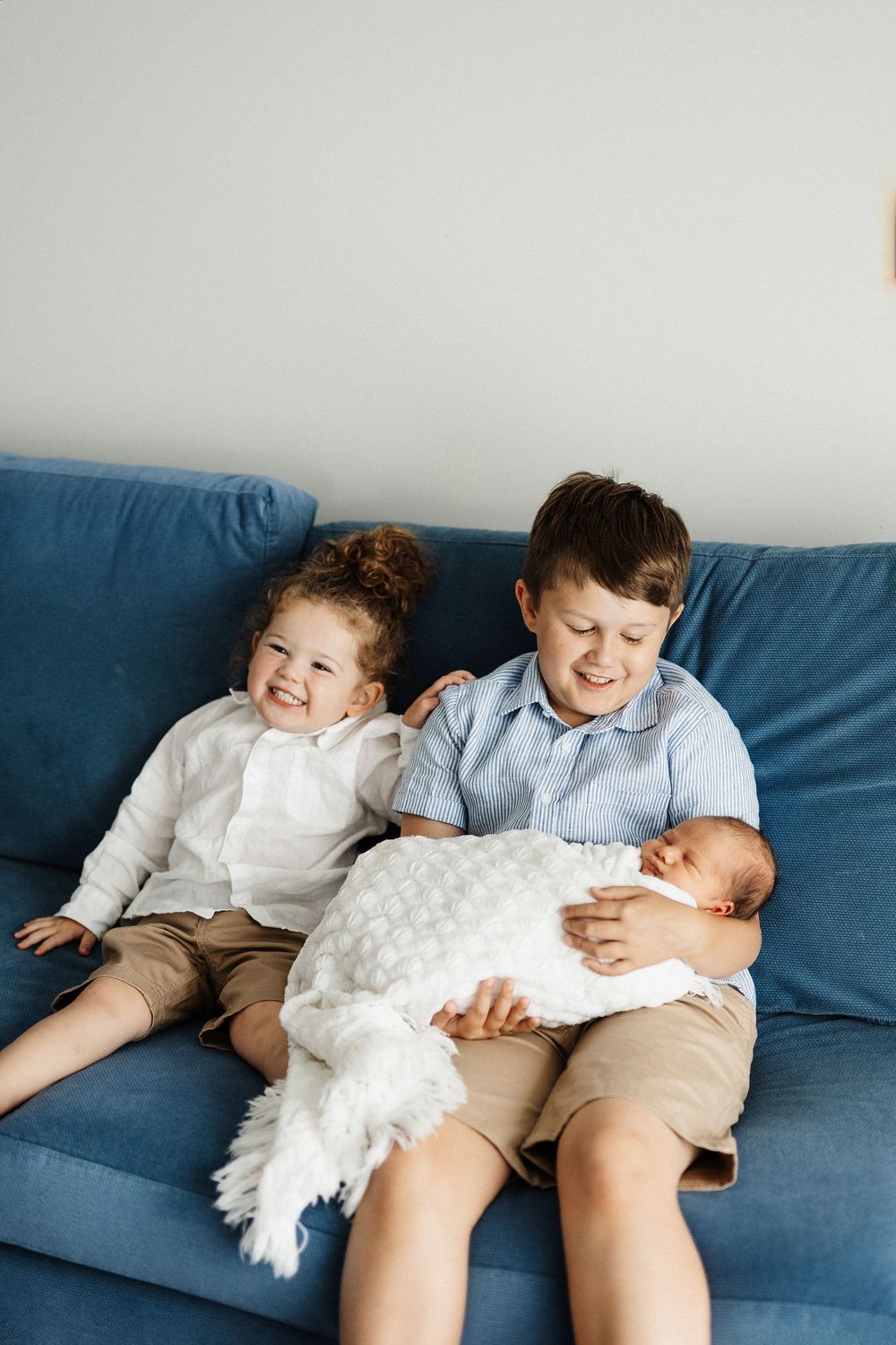 Two children sitting on a blue couch lovingly interacting with a newborn baby swaddled in white.