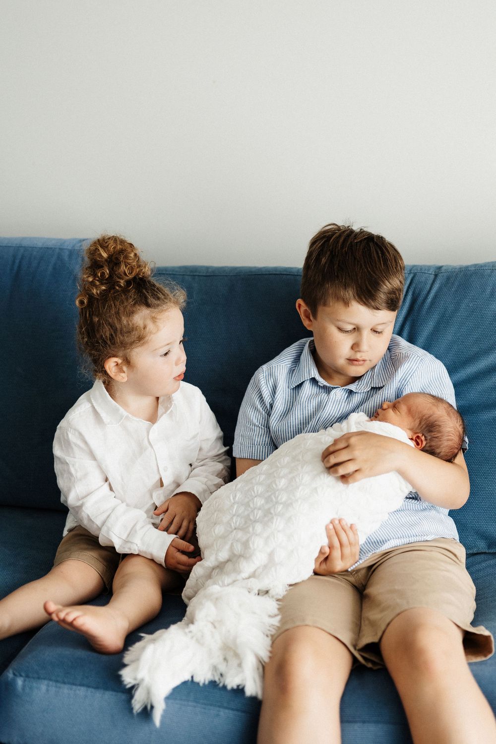 Two children sitting on a blue couch looking adoringly at a newborn baby wrapped in a white blanket.
