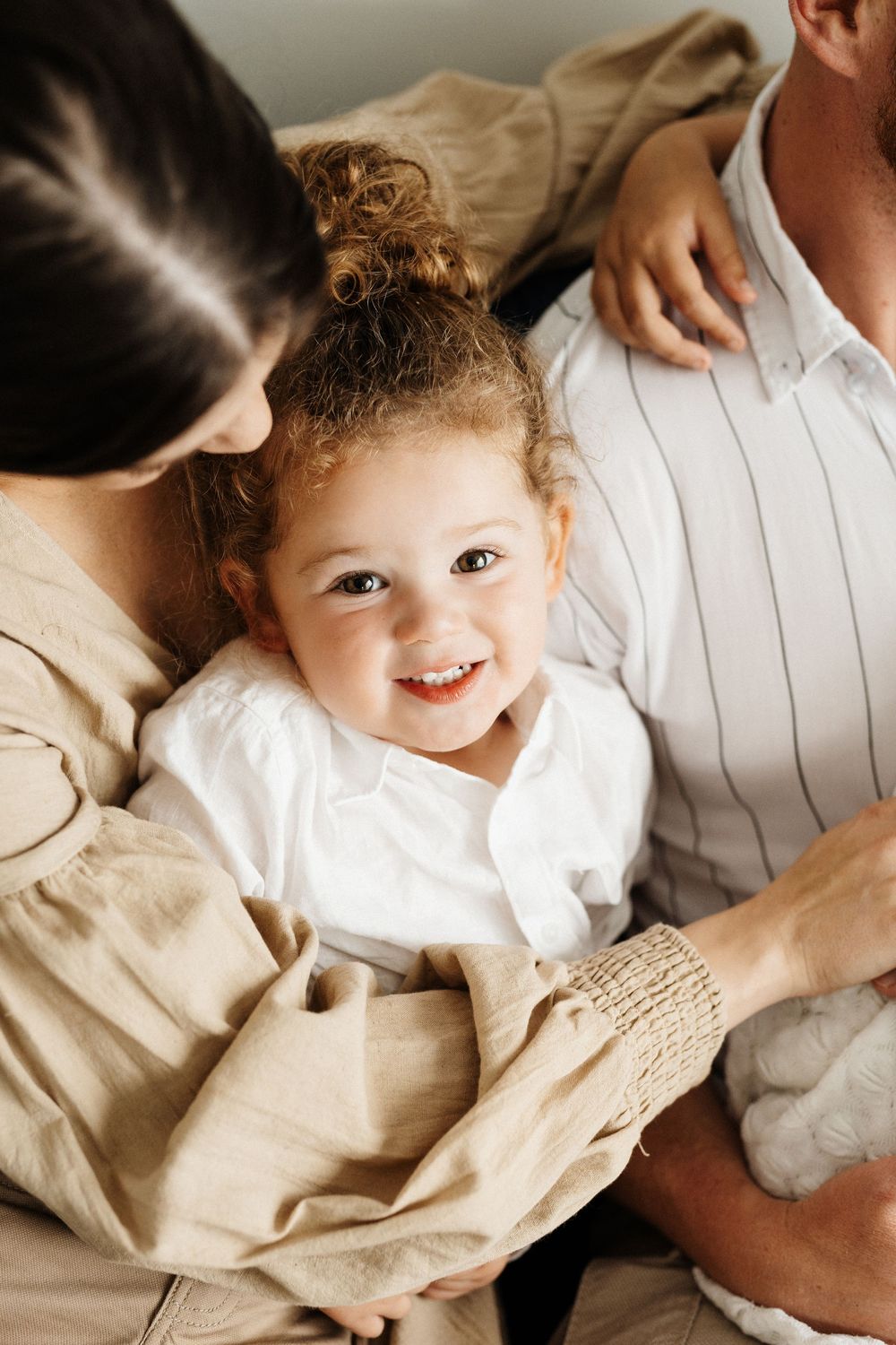 A cozy family portrait showing a small child with curly hair nestled between two adults in neutral colored clothing.