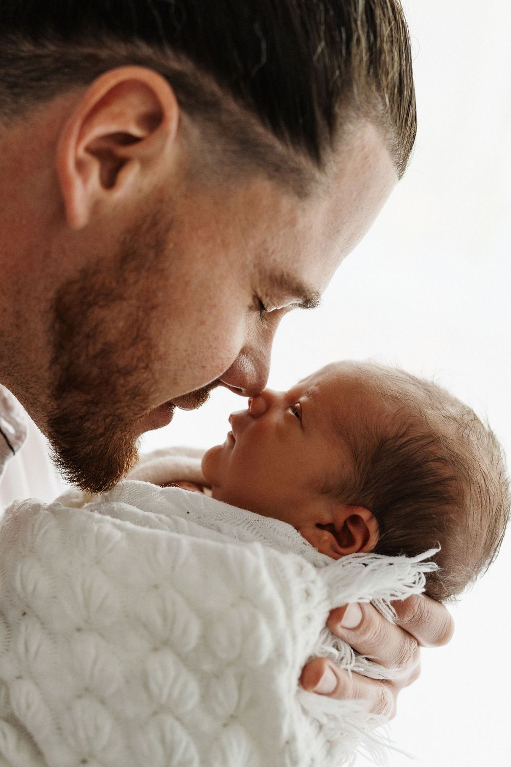 Parent tenderly bonding with sleeping newborn baby wrapped in white blanket against bright background.